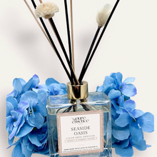 Load image into Gallery viewer, Seaside Oasis Reed Diffuser by Pure Essence Scent-A glass reed diffuser filled with scented liquid. There’s a gold cap and black/natural reeds, some featuring decorative flowers.