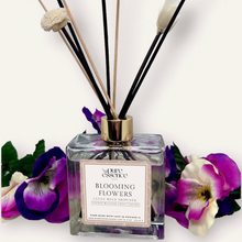 Load image into Gallery viewer, Blooming Flowers Reed Diffuser by Pure Essence Scent-A glass reed diffuser filled with scented liquid. There’s a gold cap and black/natural reeds, some featuring decorative flowers.