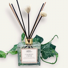 Load image into Gallery viewer, Calming Creek Reed Diffuser by Pure Essence Scent-A glass reed diffuser filled with scented liquid. There’s a gold cap and black/natural reeds, some featuring decorative flowers.