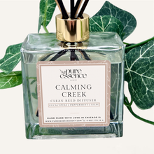 Load image into Gallery viewer, Calming Creek Reed Diffuser by Pure Essence Scent-A glass reed diffuser filled with scented liquid. There’s a gold cap and black/natural reeds, some featuring decorative flowers.