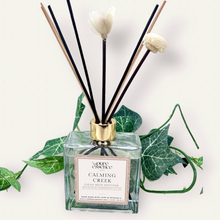 Load image into Gallery viewer, Calming Creek Reed Diffuser by Pure Essence Scent-A glass reed diffuser filled with scented liquid. There’s a gold cap and black/natural reeds, some featuring decorative flowers.