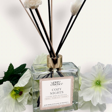 Load image into Gallery viewer, Cozy Nights Reed Diffuser by Pure Essence Scent-A glass reed diffuser filled with scented liquid. There’s a gold cap and black/natural reeds, some featuring decorative flowers.
