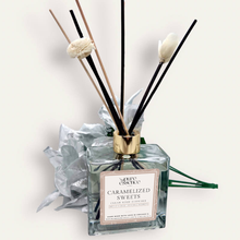 Load image into Gallery viewer, Caramelized Sweets Reed Diffuser by Pure Essence Scent-A glass reed diffuser filled with scented liquid. There’s a gold cap and black/natural reeds, some featuring decorative flowers.