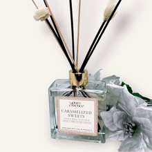 Load image into Gallery viewer, Caramelized Sweets Reed Diffuser by Pure Essence Scent-A glass reed diffuser filled with scented liquid. There’s a gold cap and black/natural reeds, some featuring decorative flowers.