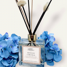 Load image into Gallery viewer, Mango Ciruela Reed Diffuser by Pure Essence Scent-A glass reed diffuser filled with scented liquid. There’s a gold cap and black/natural reeds, some featuring decorative flowers.