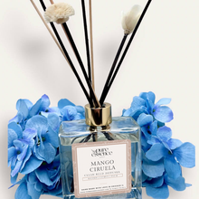 Load image into Gallery viewer, Mango Ciruela Reed Diffuser by Pure Essence Scent-A glass reed diffuser filled with scented liquid. There’s a gold cap and black/natural reeds, some featuring decorative flowers.