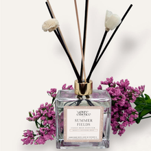 Load image into Gallery viewer, Summer Fields Reed Diffuser by Pure Essence Scent-A glass reed diffuser filled with scented liquid. There’s a gold cap and black/natural reeds, some featuring decorative flowers.